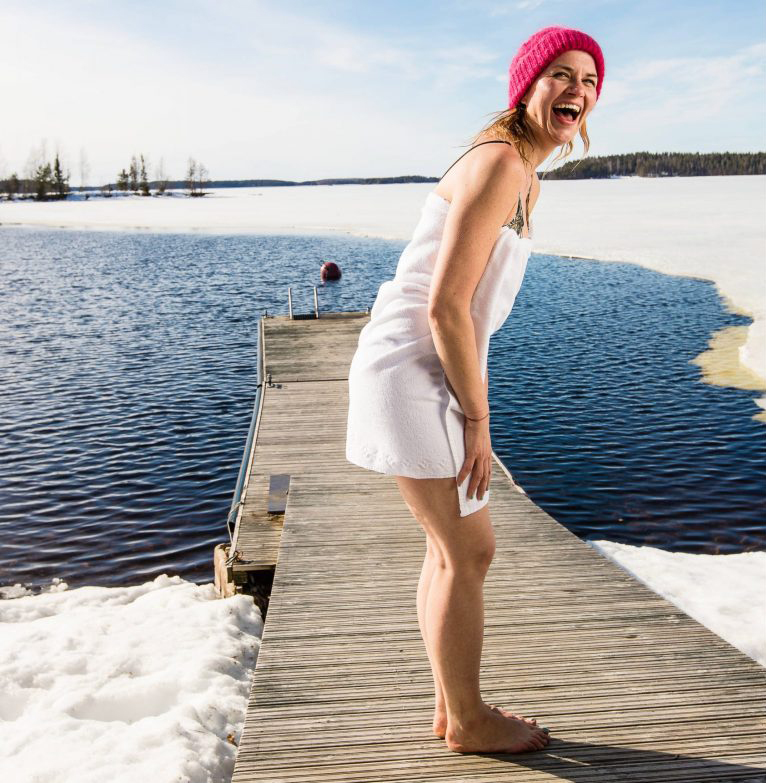 A smiling woman in a white towel and pink snow hat stands on a wooden dock by a frozen lake. 