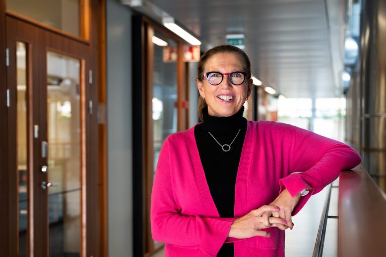 Katriina Aalto-Setälä looks into the camera and smiles. She wears a bright pink sweater over a black turtleneck with matching pink and black glasses.