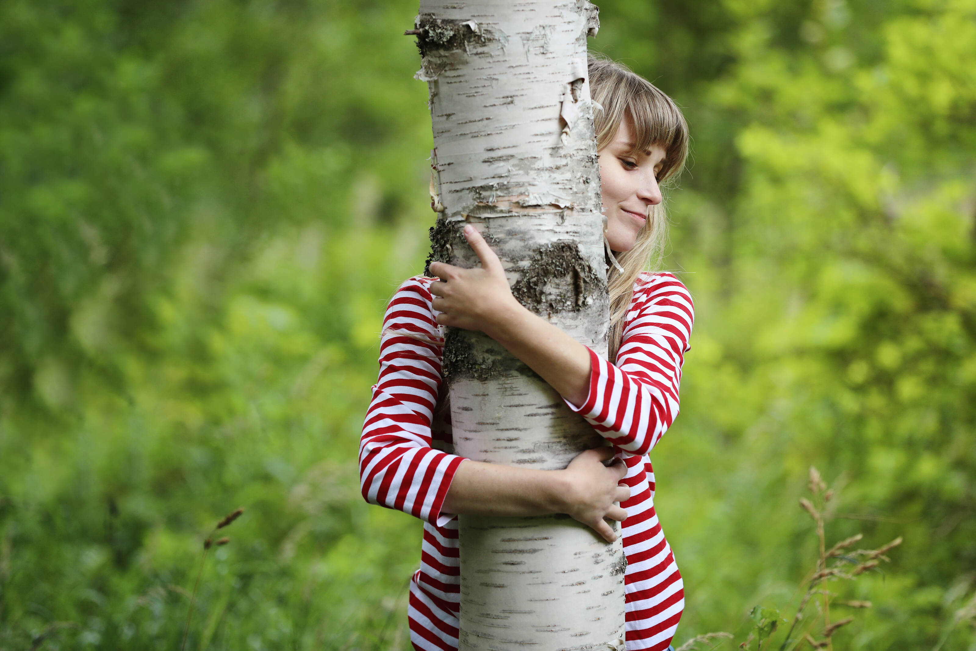 A blonde woman in a red and white striped shirt hugs a birch tree.