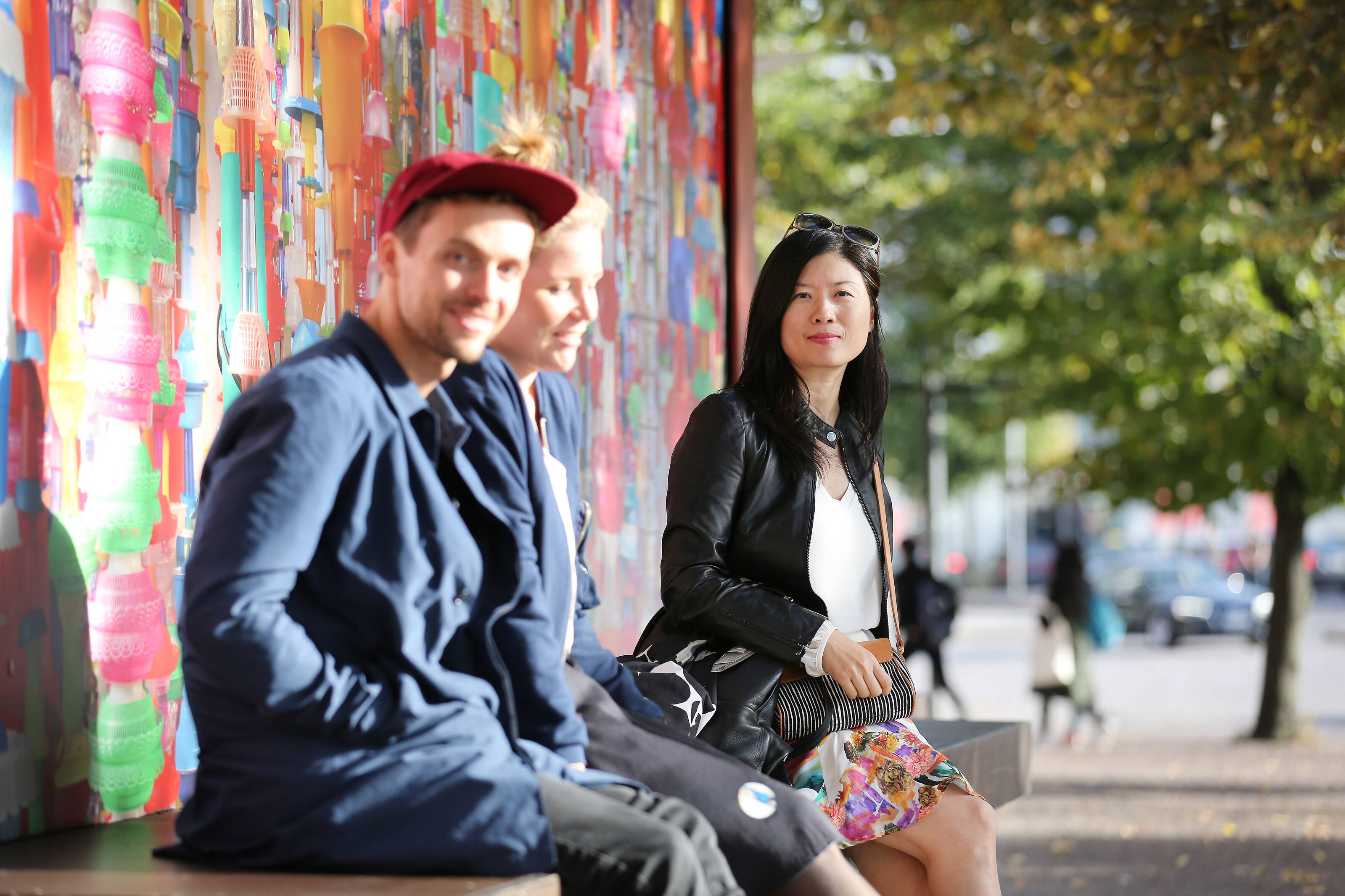 A man and two women sit outside in the sunshine by a colourful mural.