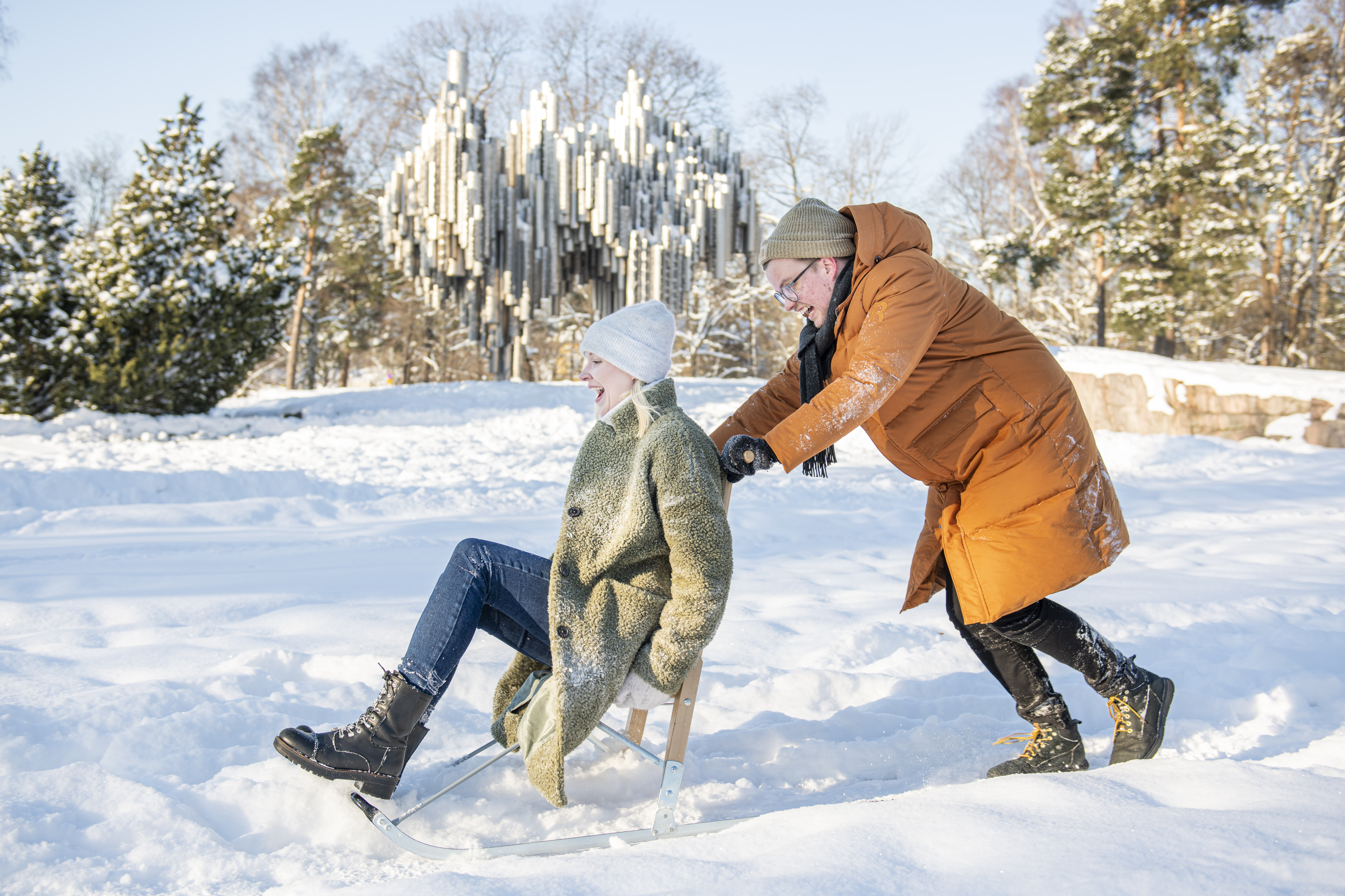 A man in an orange jacket and green cap pushes a laughing woman in jeans, boots, a green jacket and a white cap through the snow on a kick sled. 