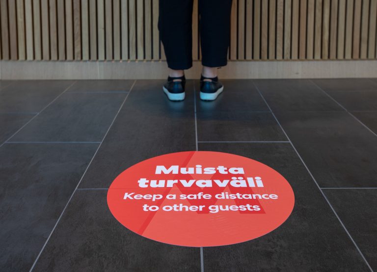 The legs of a person wearing black pants can be seen behind a red sign painted on the floor of the Messukeskus centre that reads “Keep a safe distance to other guests” in both English and Finnish.