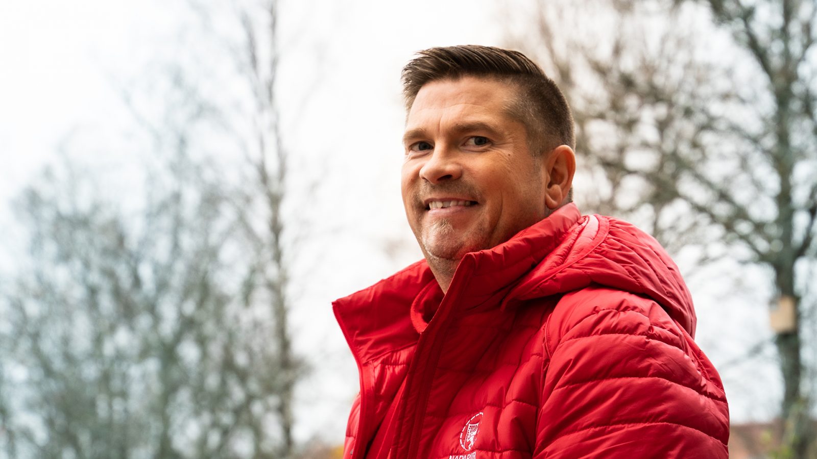 Man in a red shirt and red jacket smiling towards the camera with trees in the background.
