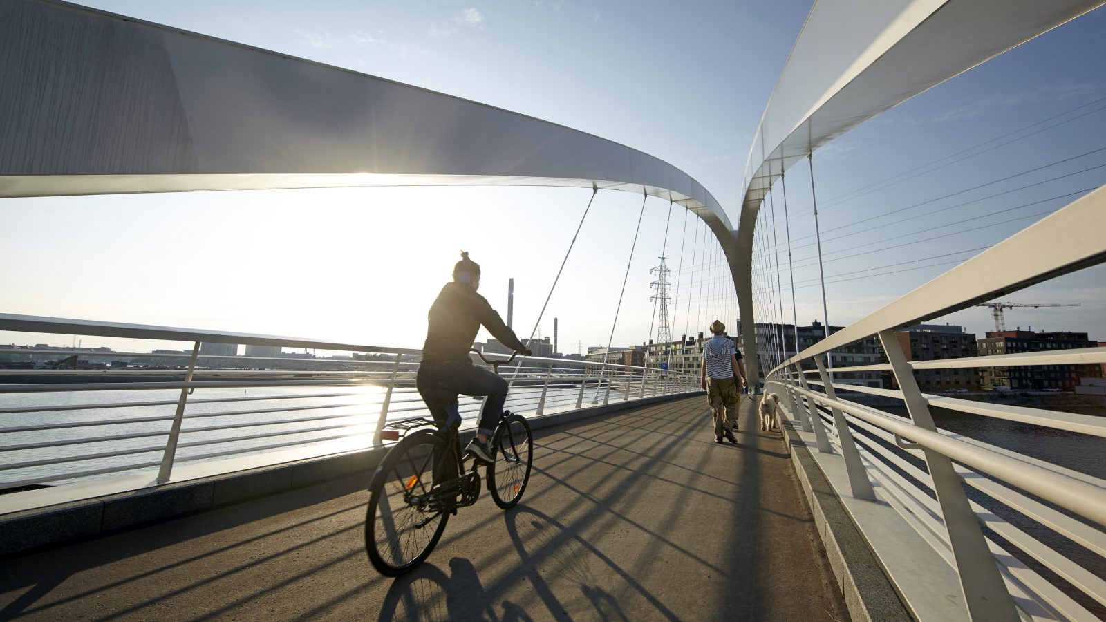 A woman biking whilst a couple are walking a dog across a bridge on a sunny day in Helsinki Finland.