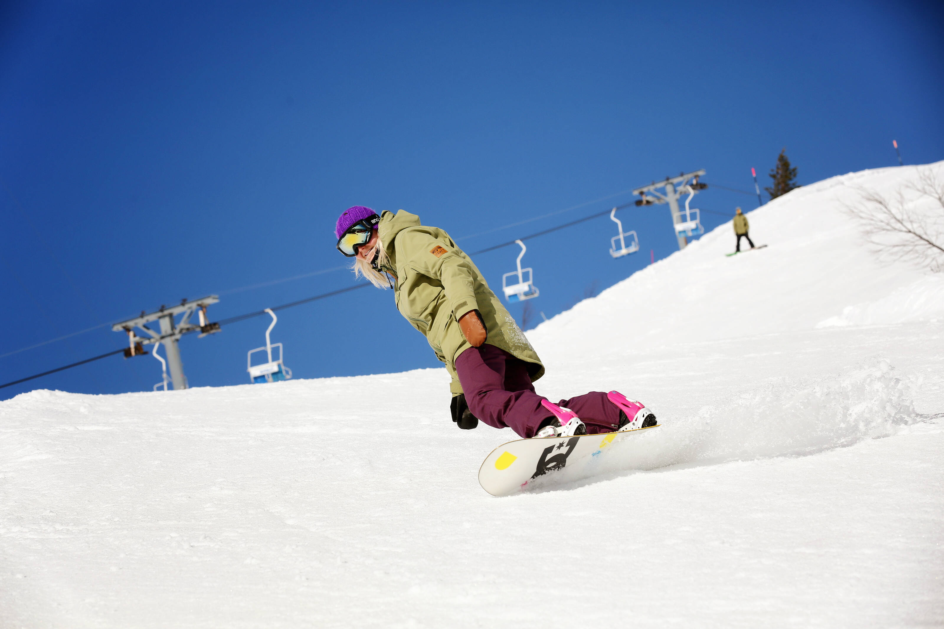 A female snowboarder in a green jacket, purple cap and ski goggles carves down a ski slope. There's a ski lift in the background.