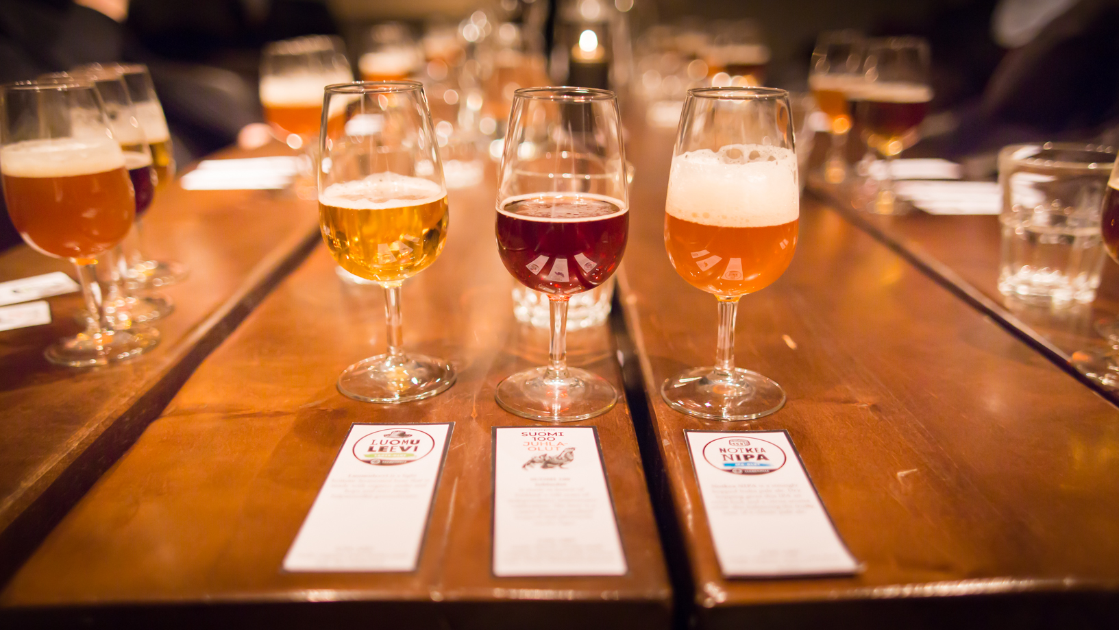A flight of three beers in long-stemmed glasses sits at eye level on a wooden table.