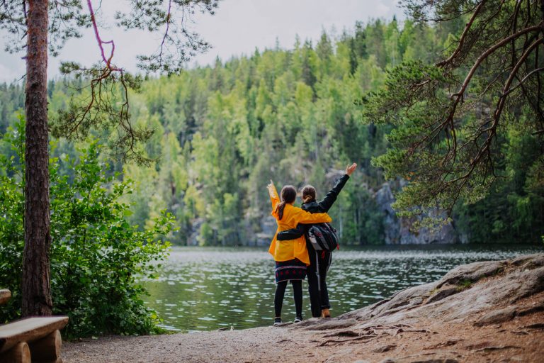 Two women with one of their arms around one another and the other arm in the air stand with their backs to the camera by a Finnish lake. 
