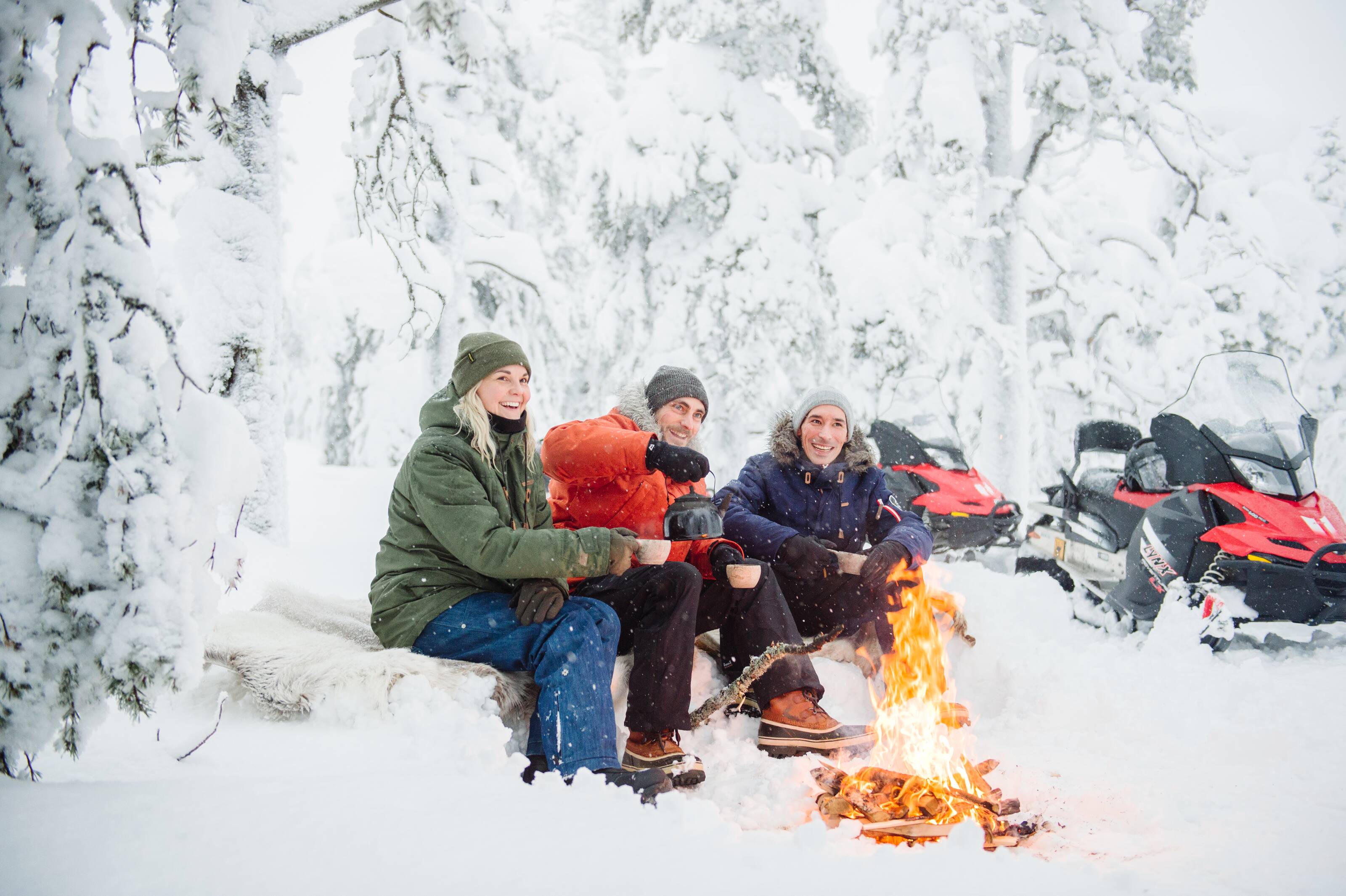 Three people sitting by a fire in a snow-covered forest