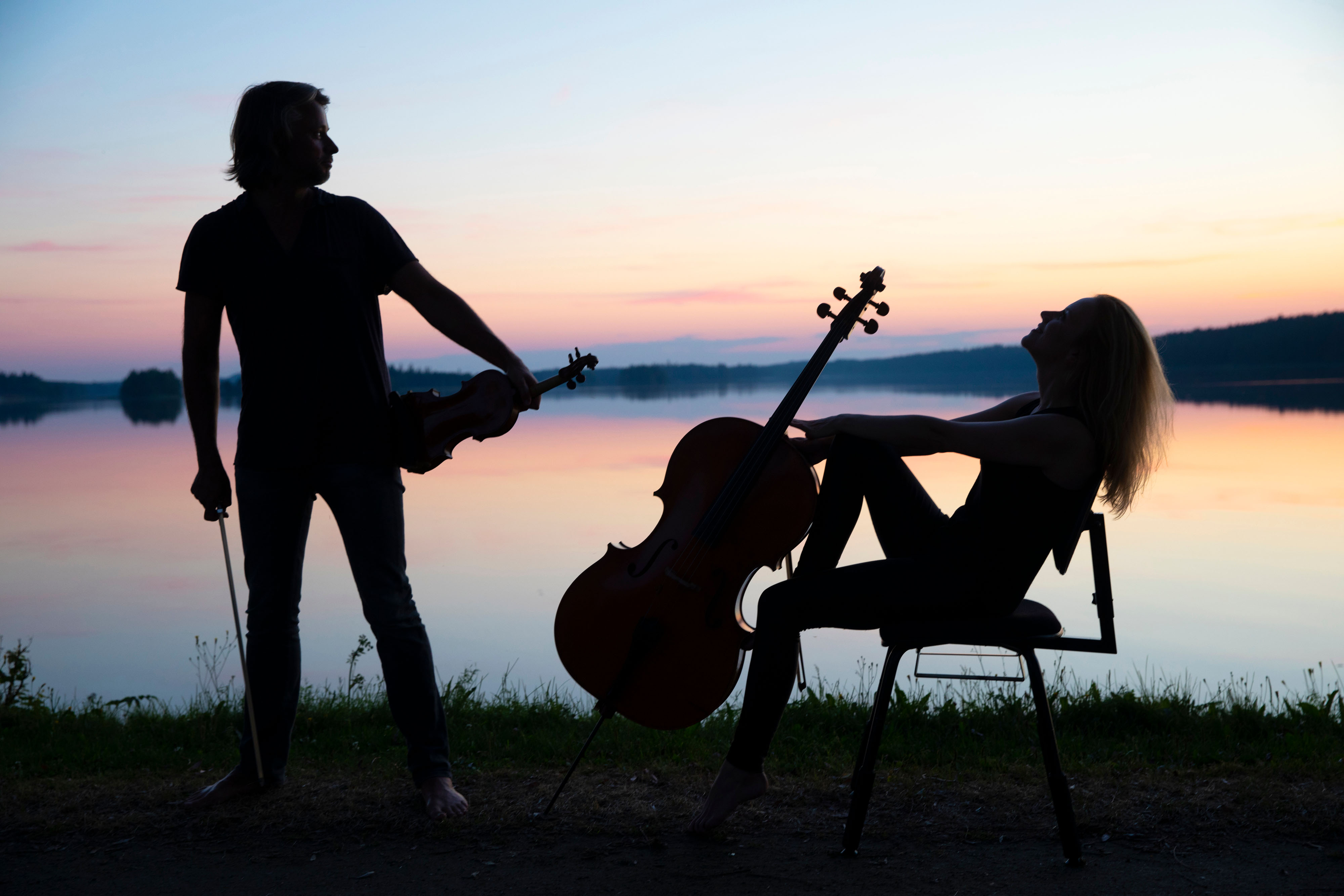A man holding a violin and a woman with a cello are silhoueted by the sunset next to a lake.