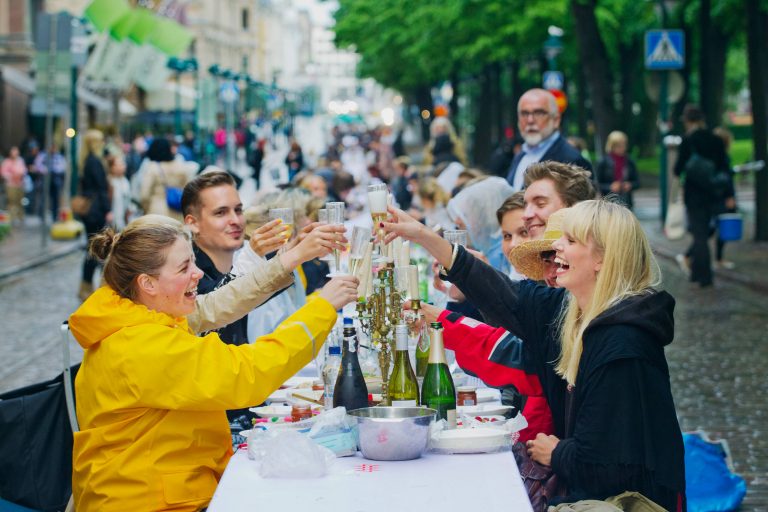 A group of friends celebrating and cheering with champagne glasses by an outdoor table in the middle of the street