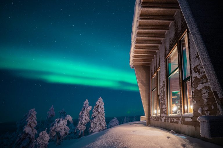The Northern Lights glow greenish-blue over a wooden building and snow-covered forest at the ultra-luxury Octola Private Wilderness resort.