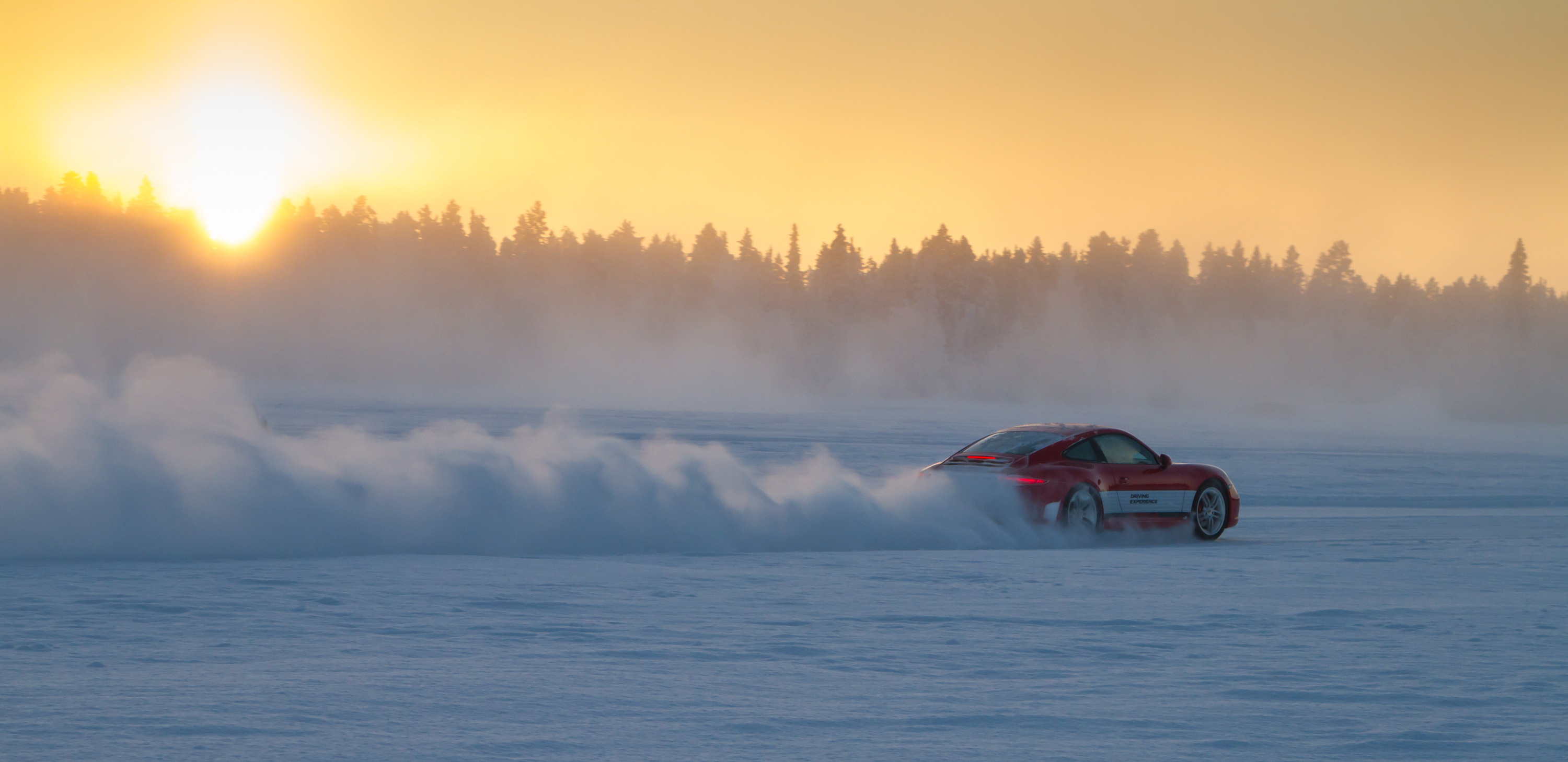 A red Porsche with a white stripe speeds along a snowy trail near sunset, leaving a cloud of snow in its wake.