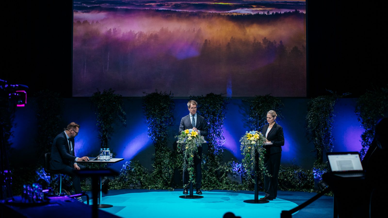 Three people dressed in dark business suits on a stage with flowers and green plants.