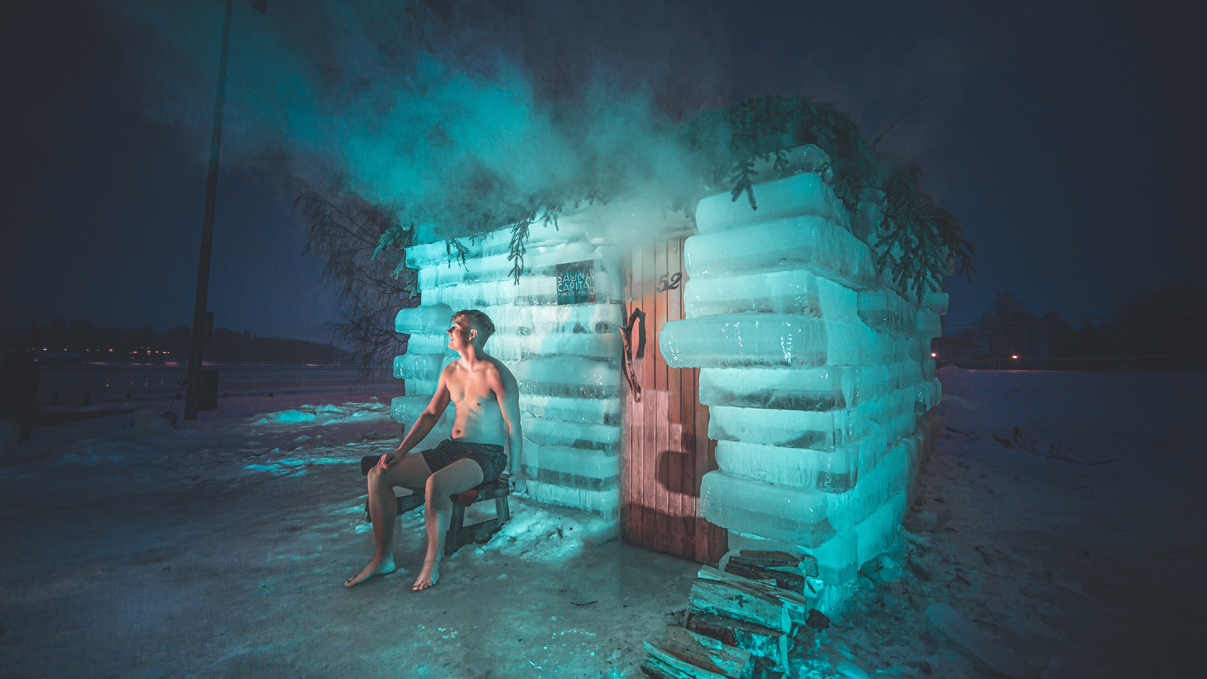 A shirtless man sits on a wooden chair outside a steaming sauna made of ice at nighttime.